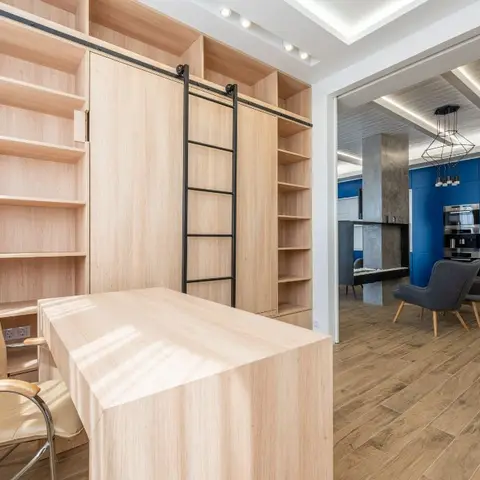 Built-in light-oak study wall with a rolling library ladder and matching desk, opening through to a blue-cabinet kitchen and lounge beyond.