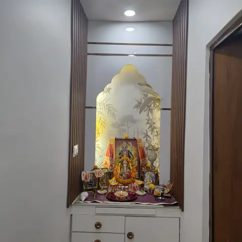 Built-in pooja niche framed by fluted wood columns with a backlit arched botanical glass panel, idols dressed in marigold garlands
