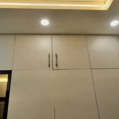 Built-in pooja niche with a lit glass-front display housing a Ganesha idol, set within a run of cream wardrobe cabinetry.