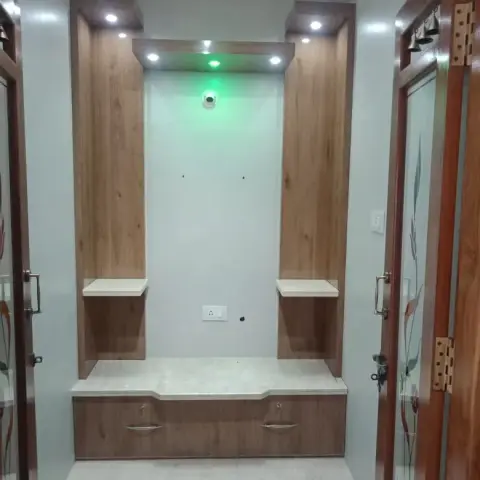 Built-in pooja unit niche with wood-veneer twin towers, a marble seating ledge, storage drawers below and a green accent light illuminating the shrine recess.