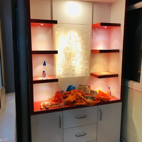 Built-in pooja unit with a backlit deity relief panel, open display shelves lit in red-orange accent lighting, and grey laminate storage drawers below.