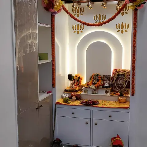 Built-in pooja unit with a backlit arched niche framed by gold lotus motifs, marigold garlands and bells, over a lower cabinet with drawers.