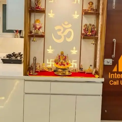 Built-in pooja unit with a backlit CNC-cut Om and swastik panel, deity idols on hanging shelves, set into a white cabinetry niche beside the kitchen.