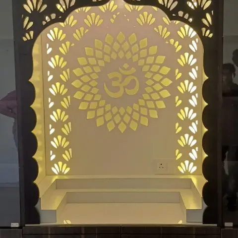 Built-in pooja unit with a backlit laser-cut arch jaali panel centred on an Om motif, set into a dark wood-grain cabinet with drawer storage below.