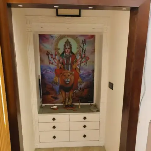 Built-in pooja unit with a carved cream cabinet niche housing a deity poster and idols above a drawer console.