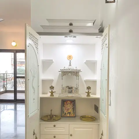 Built-in pooja unit with arched frosted-glass cabinet doors, hanging brass bells, a carved silver temple, and brass ritual items on a marble-effect shelf.