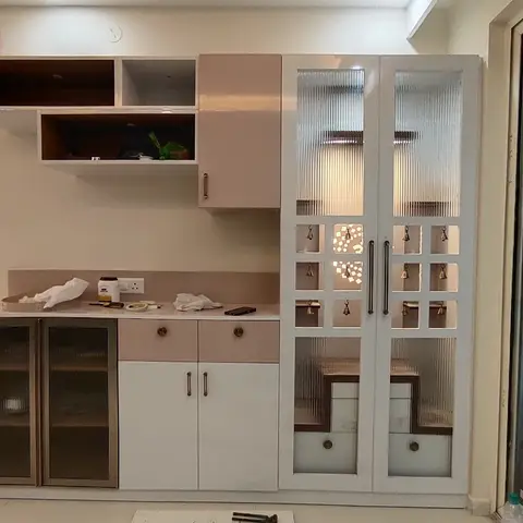 Built-in pooja unit with fluted glass doors and hanging brass bells beside a blush-and-white crockery storage unit, in an apartment with a balcony view.