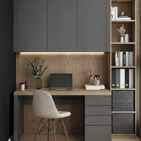 Built-in study nook with a wood desk under charcoal overhead cabinets, a backlit wood panel, open bookshelf, and a cream Eames-style chair.