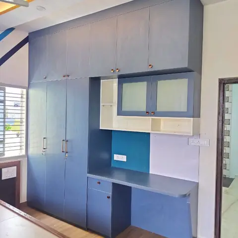 Built-in wardrobe and study unit in slate-blue laminate with glass-shutter overhead cabinets and a rounded-corner study desk, wood-tone flooring