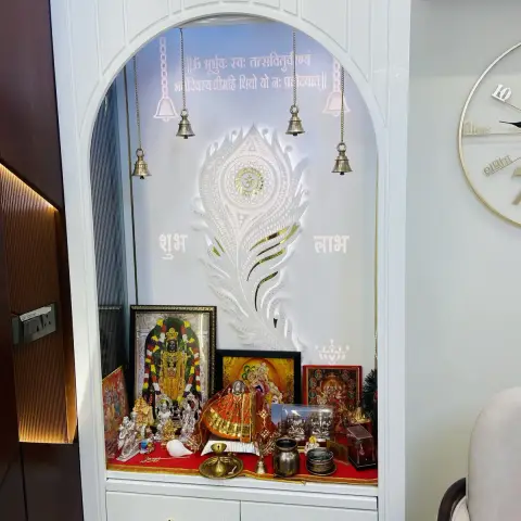 Built-in white lacquered pooja unit with an arched backlit niche, a peacock-feather motif panel, hanging bells, and idols set on a red cloth.