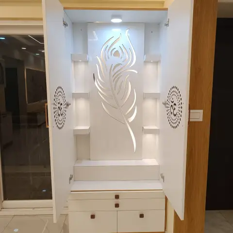 Built-in white mandir unit photographed with its shutters swung open, showing a backlit CNC peacock-feather panel between stepped display ledges