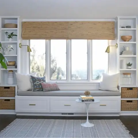 Built-in window seat nook with bamboo roman shades, brass reading sconces, flanking bookshelves with woven storage baskets and a round pedestal side table.
