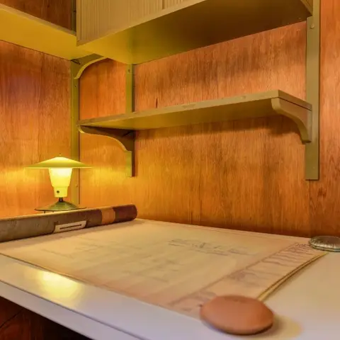 Built-in wood-panelled study nook with a floating desk, bracket shelf, and a retro mushroom-shaped table lamp lighting a rolled blueprint.