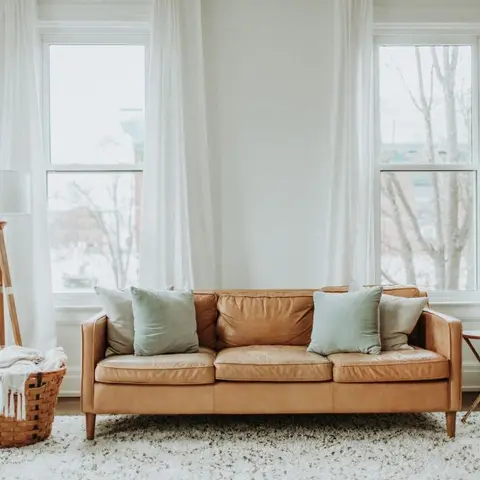 Camel leather sofa with sage cushions against tall sheer-curtained windows, a woven basket and tripod floor lamp beside a shag rug.