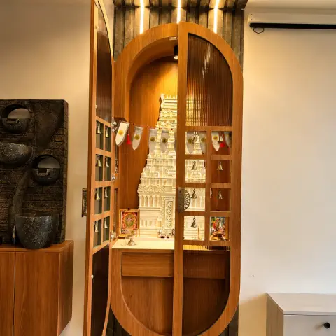 Capsule-arch teak mandir whose fluted glass doors open on rows of brass bells to a white marble gopuram in a backlit niche