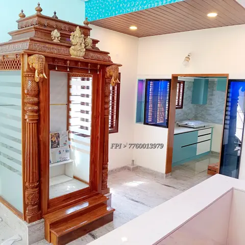 Carved teak pooja mandir with elephant-motif pillars and frosted glass shutters, set in a marble-floored alcove next to an open teal-cabinet kitchen.