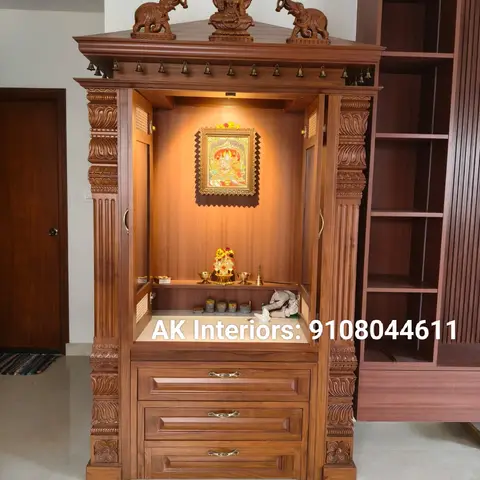 Carved teak pooja unit with a temple-style pitched roof topped by elephant and goddess figures, fluted pillars, brass bells along the cornice
