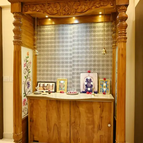 Carved teak pooja unit with temple-style pillared canopy, marble platform and hanging brass bells against a patterned metallic backdrop