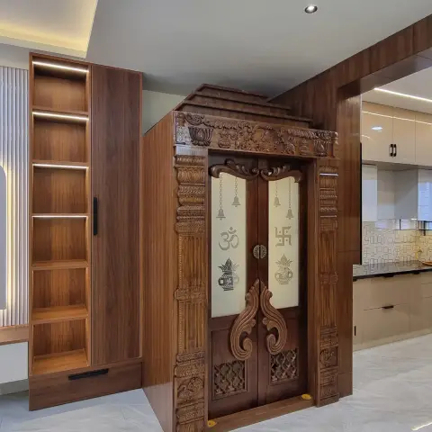 Carved wooden pooja unit with ornately detailed double doors featuring Om and Swastik motifs and hanging bells, flanked by an open wood shelving column
