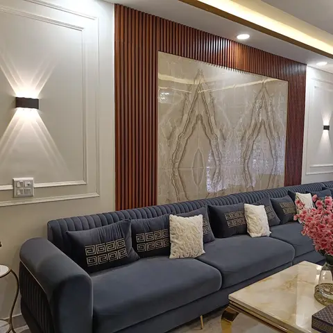 Channel-tufted navy velvet sectional facing a marble feature wall framed by fluted wood battens, lit by twin uplight-downlight wall sconces.