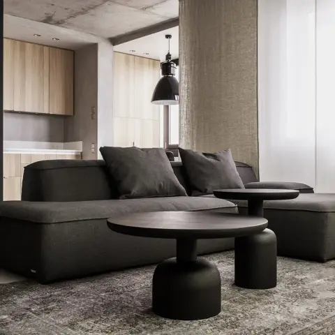Charcoal modular sofa with a deep chaise on a faded grey rug under a bare concrete beam, two black pedestal tables in front of it