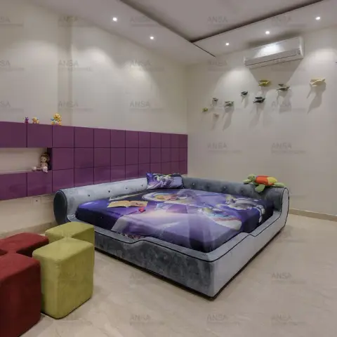 Child's room with a low grey upholstered bed under a cartoon-print quilt, a long aubergine gloss storage band with an open shelf of toys