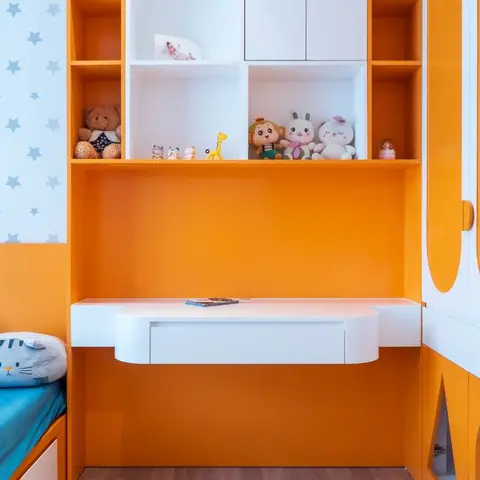 Children's bedroom with bright orange and white joinery, a floating study desk, and an open display shelf styled with plush toys.