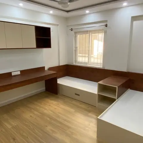 Children's bedroom with twin built-in platform beds along two walls, a matching study desk, and an overhead storage cabinet, in maroon and cream laminate.