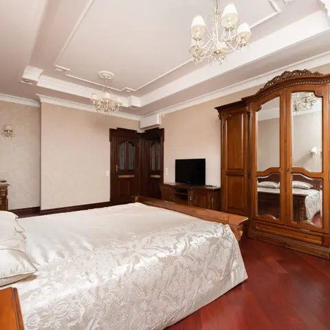 Classic bedroom with a carved wood mirrored wardrobe and matching headboard, crystal chandelier, cove-lit tray ceiling, and cherry-toned wood flooring.