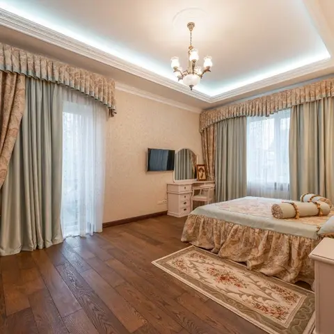 Classic bedroom with sage-green damask curtains, a crystal chandelier under a cove-lit ceiling, dark wood flooring, and a vanity dresser with an oval mirror.