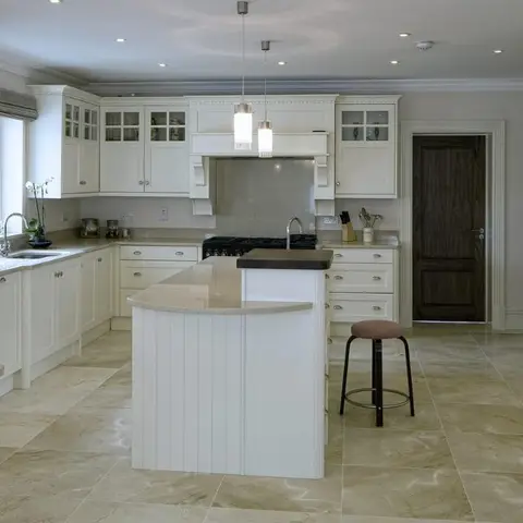 Classic cream shaker kitchen with a curved island, glass-front display cabinets, twin pendant lights over the hob, and large veined marble floor tiles.