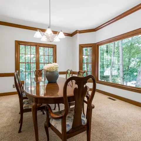 Classic dining room with an oval wood table and Queen Anne chairs beneath a brass chandelier, wood-trimmed bay windows overlooking trees
