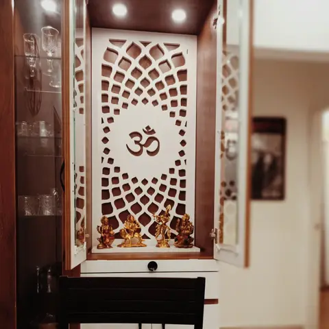 Close-up of a built-in pooja cabinet with a backlit CNC Om jaali panel and brass deity idols, flanked by a glass display cabinet.