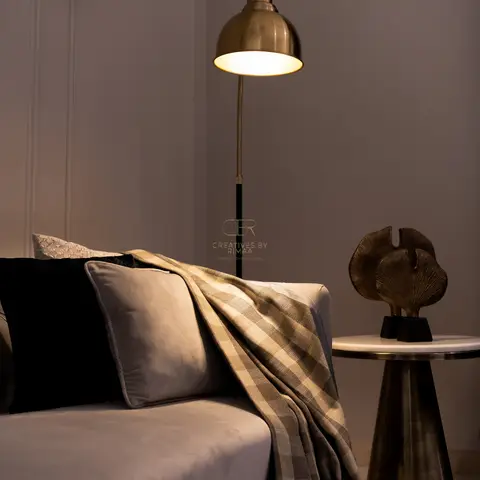 Close-up of a grey velvet sofa with a plaid throw beside a brass dome floor lamp and a marble-top side table styled with a bronze sculpture