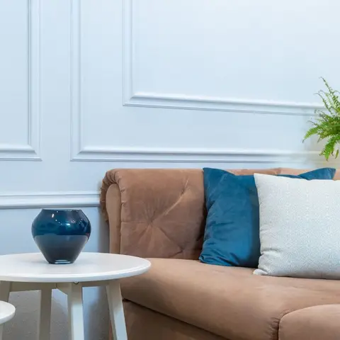 Close-up of a tan tufted sofa with teal and white cushions beside a white panel-moulding wall, a round side table, and a potted fern