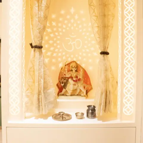 Close-up of a white pooja unit niche with a carved jaali arch, sheer gold tie-back curtains, and a backlit star-and-Om projection behind a Durga idol.