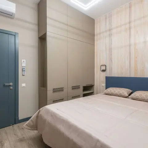 Compact bedroom with a pale pine plank-clad wall behind a navy upholstered headboard, a taupe matt wardrobe with a drawer bank and open niche