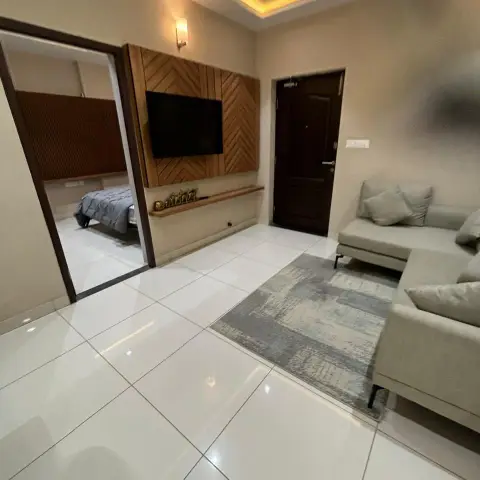 Compact flat living room where the TV sits on a chevron-laid teak panel with a slim brass-idol display ledge below, a grey L-sofa on a distressed grey rug