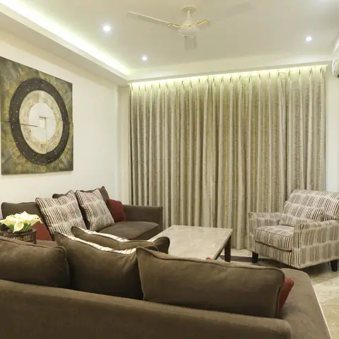 Compact Indian living room with brown fabric sofas around a marble coffee table beneath a cove-lit ceiling and floor-length curtains.