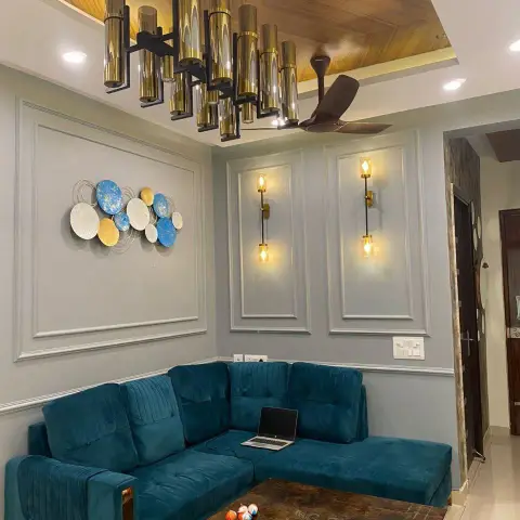 Compact living room photographed at night: a teal velvet L-sofa with a laptop on it, grey walls carved into white moulding frames with brass tube sconces