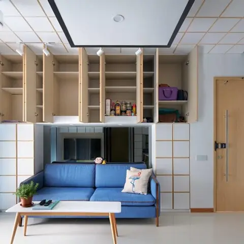 Compact living room with a blue sofa and white coffee table beneath open overhead storage cabinets in light wood, gridded false ceiling with track lighting