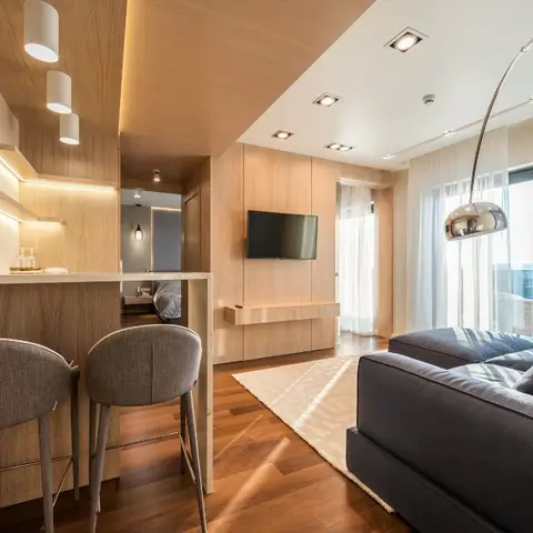 Compact living room with a wood-panelled TV wall, an arc floor lamp beside a charcoal sofa, and a wood breakfast bar with grey stools opening into the kitchen.