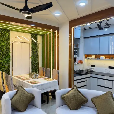 Compact open-plan flat seen over a white curved sofa: a lace-covered dining table backed by strips of artificial green-leaf wall inside nested gold LED reveal