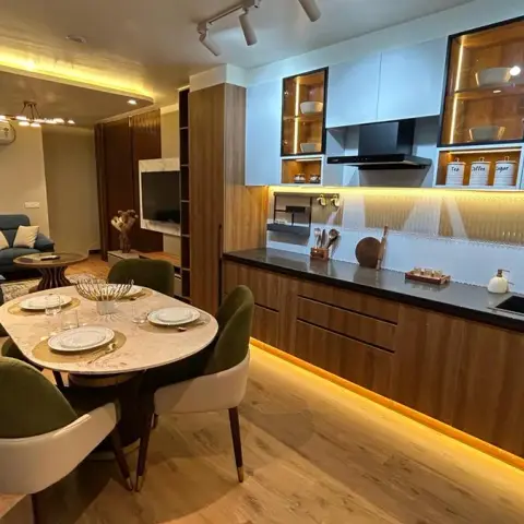 Compact open-plan flat where a straight-line kitchen in walnut base units and powder-blue matt wall cabinets faces a round marble-top dining table with olive