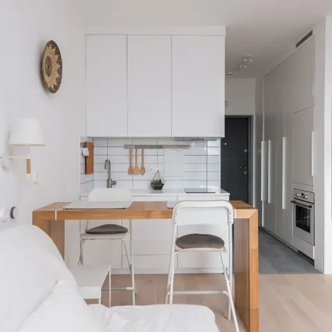 Compact open-plan studio with a white subway-tile kitchenette, a wood breakfast bar with stools, and a white sofa in the foreground