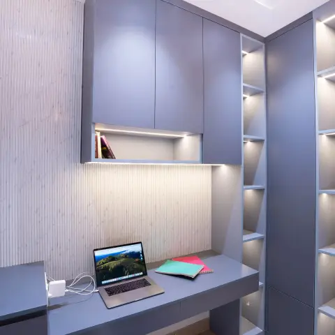 Compact study nook with a grey built-in desk under fluted-panel-backed wall cabinets and a tall backlit open shelving tower.