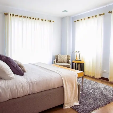 Corner bedroom wrapped on two sides by cream eyelet sheers on brass rods, with a plum bolster on a white duvet