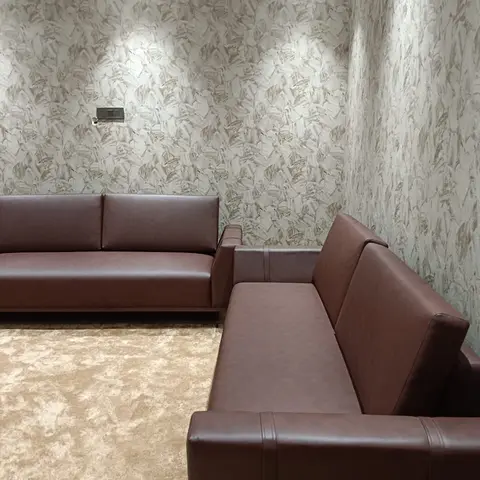 Corner living room seating with a brown leatherette L-shaped sofa against a textured beige wallpapered wall and a plush cream rug.