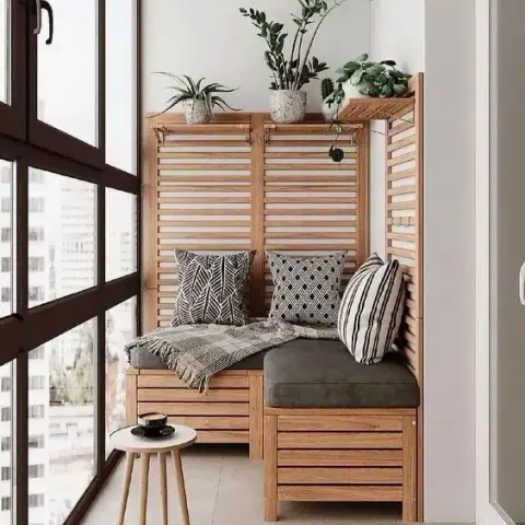 Corner of a glazed high-rise balcony with an L-shaped slatted-wood storage bench, black-and-white patterned cushions and potted greens lined along its shelf