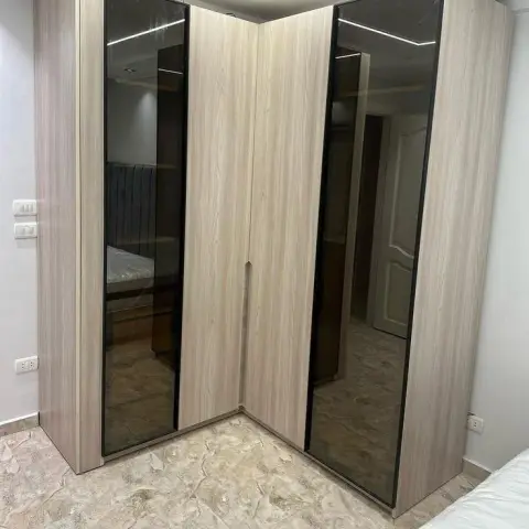 Corner-return wardrobe in pale ash laminate with full-height bronze-tinted mirror strips on its shutters